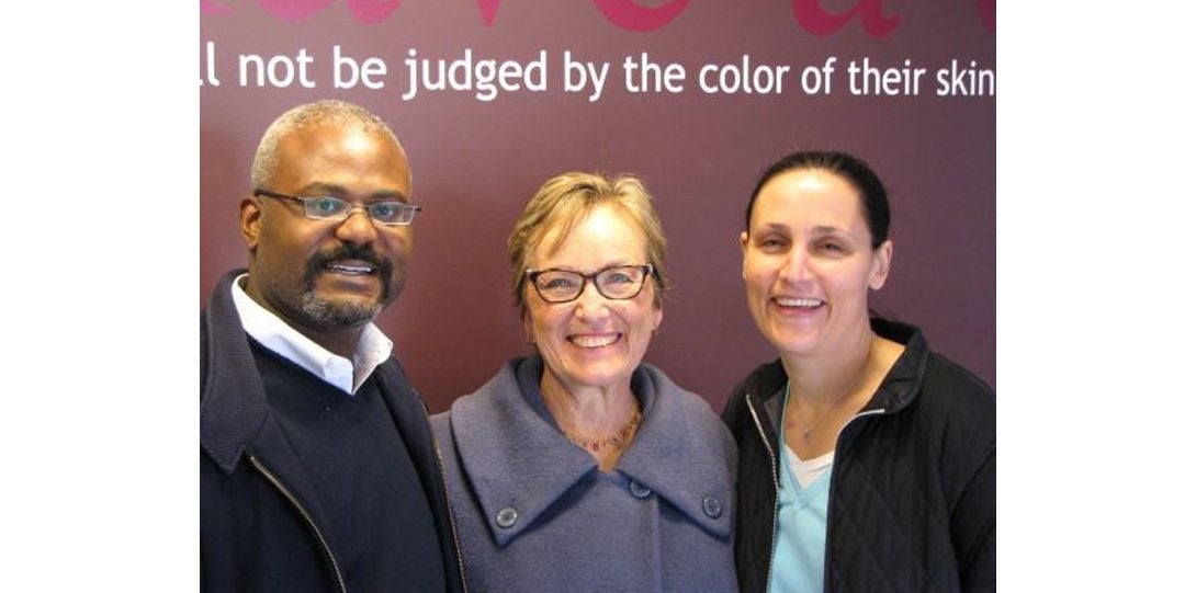 three people posing for photograph in front of a wall with partial phrase stating 