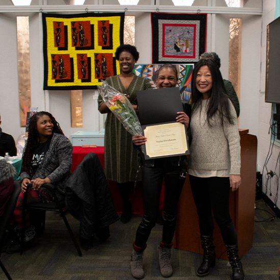 student holding flowers and award certificate posing with professor as others look on