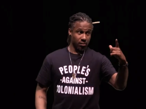 A person in a black shirt that reads Peoples against Colonialism is photographed while speaking in a lecture