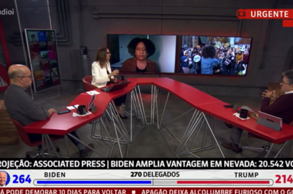 A newsroom with three people interview a person featured on screen