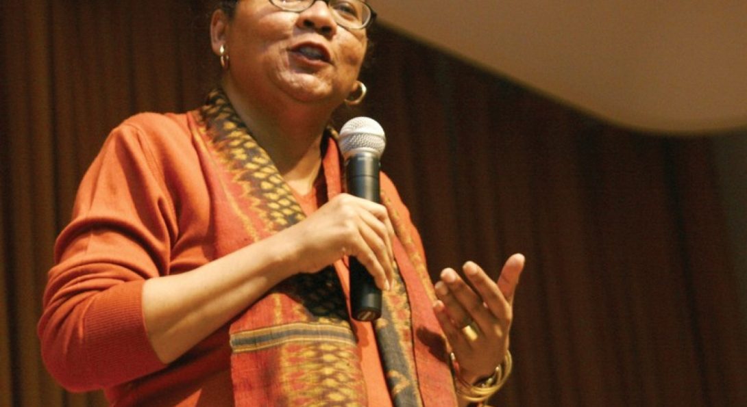black woman with black glasses wearing orange top with printed scarf speaking into a microphone