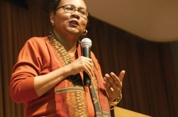 black woman with black glasses wearing orange top with printed scarf speaking into a microphone