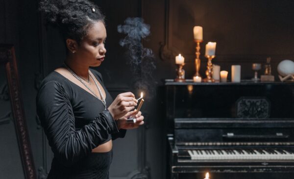 Black woman standing at the edge of the photo lighting up a piece of wood. the brackground shows a half of a piano and multiple candles lighten up on top of it.