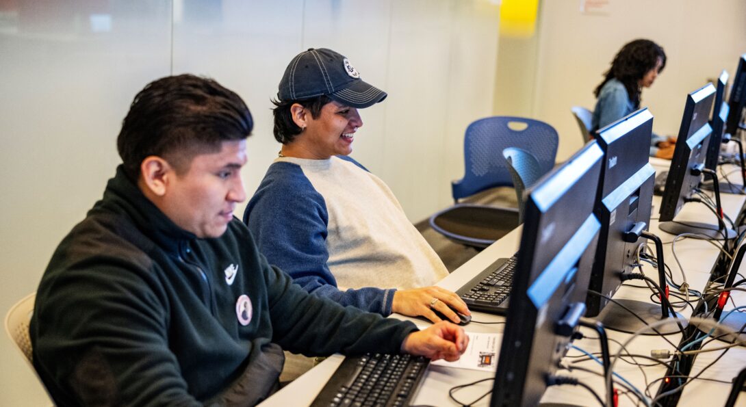 three students working at a row of computers. one student has a jean jacket, and another student is wearing a baseball cap and smiling. the last student wears a jacket with at Douglass Day pin