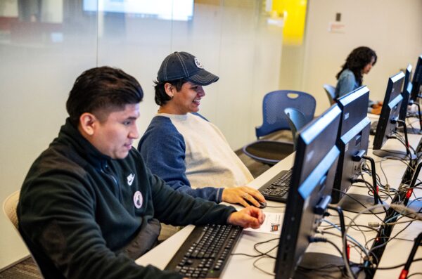 three students working at a row of computers. one student has a jean jacket, and another student is wearing a baseball cap and smiling. the last student wears a jacket with at Douglass Day pin