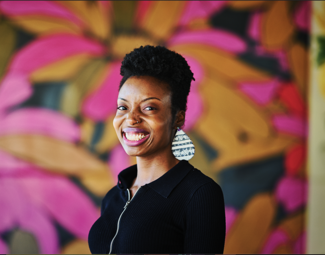 woman wearing a black jacket and white patterned earring smiling at the camera standing against a floral patterned background