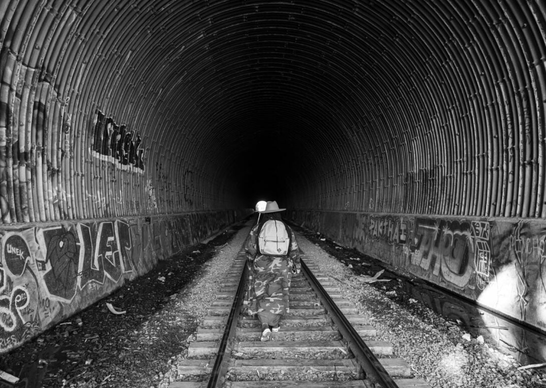 black and white photo. person wearing camo and a backpack walking on railroad tracks in a large tunnel