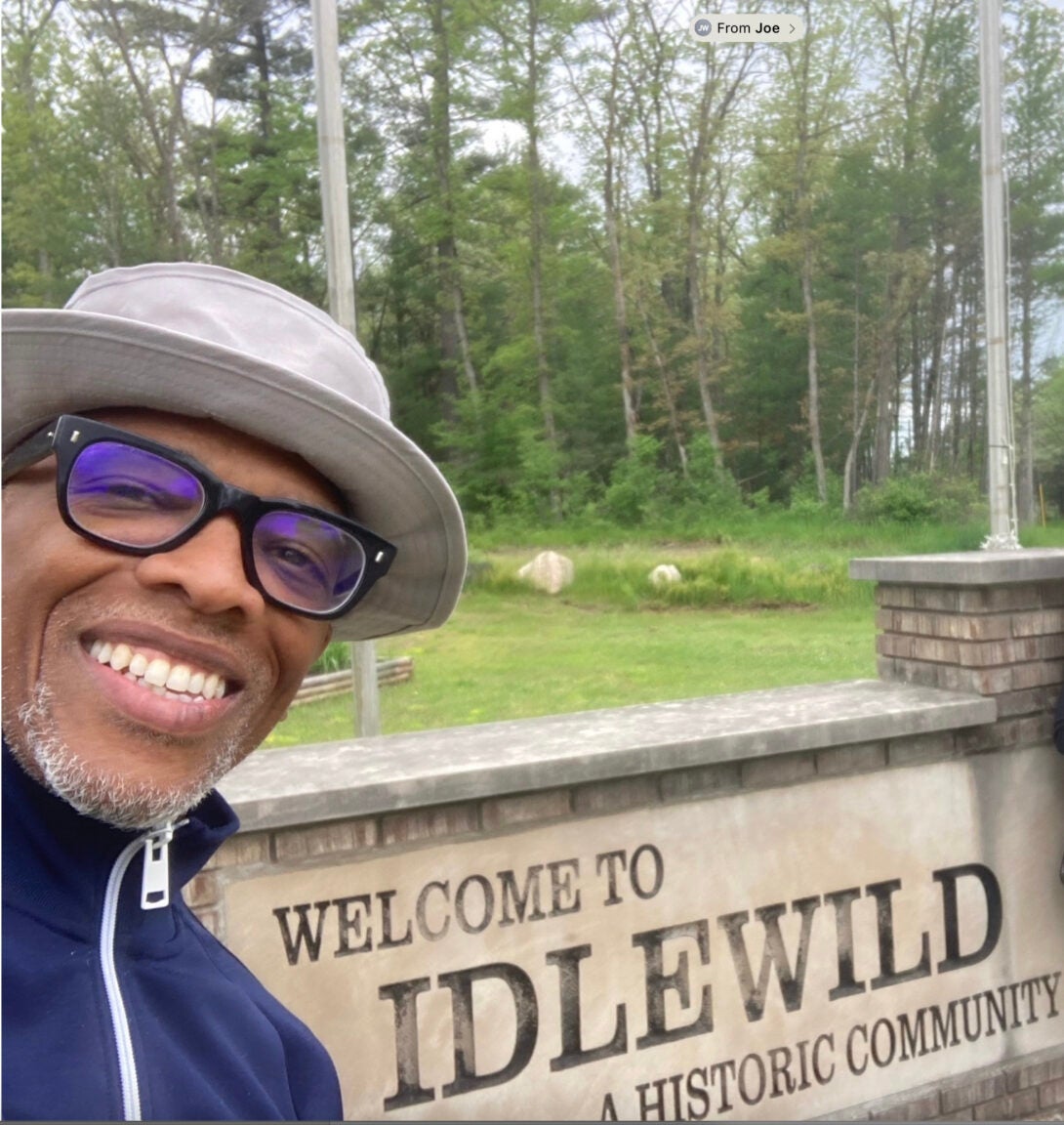 person wearing a hat and glasses smiling at a camera next to a sign that says 