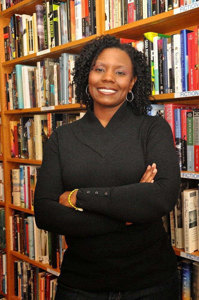 person wearing a black suit smiling and standing in front of book cases